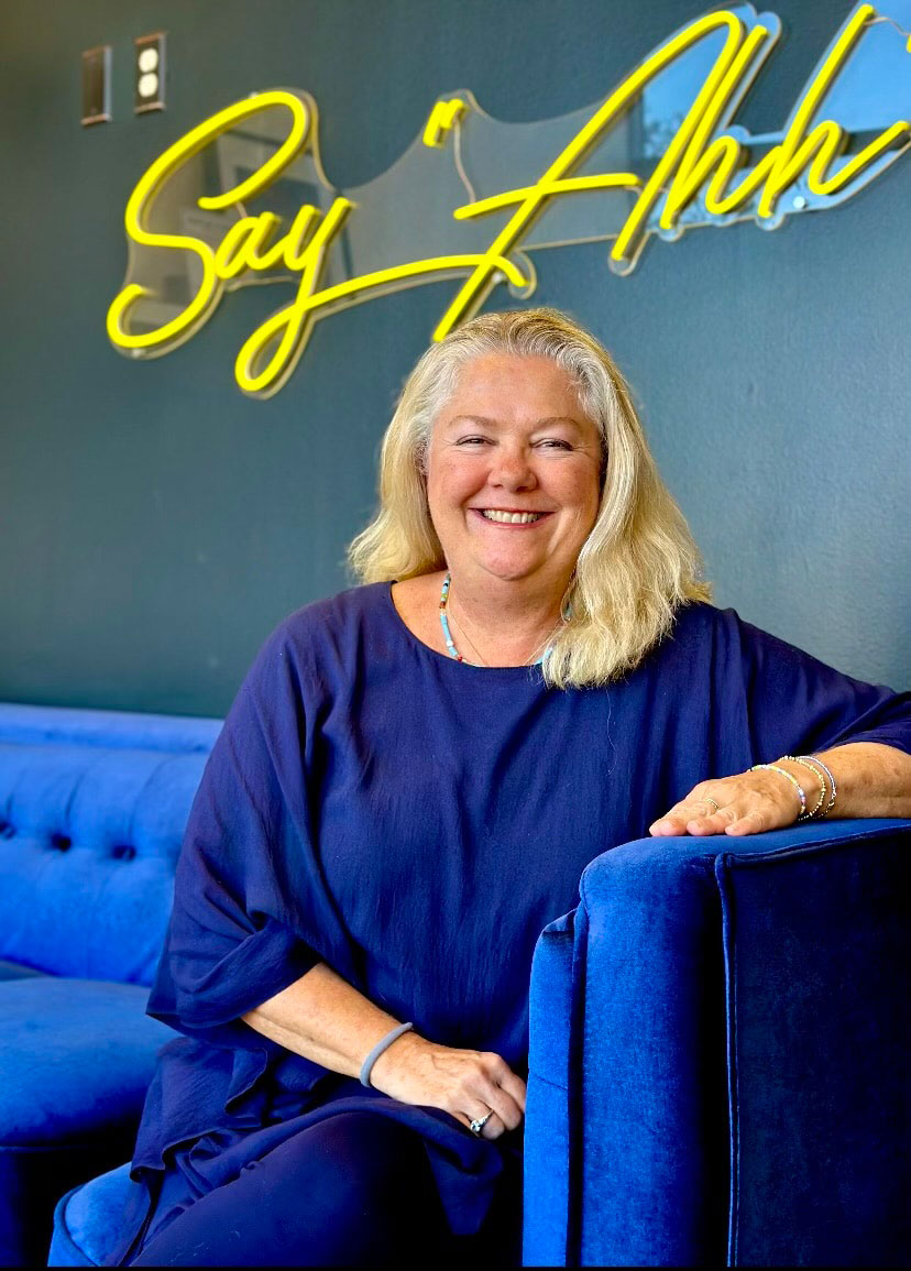 A woman with blonde hair sits in front of a blue sofa, smiling at the camera, positioned in front of a sign with yellow text reading  SAY HI .