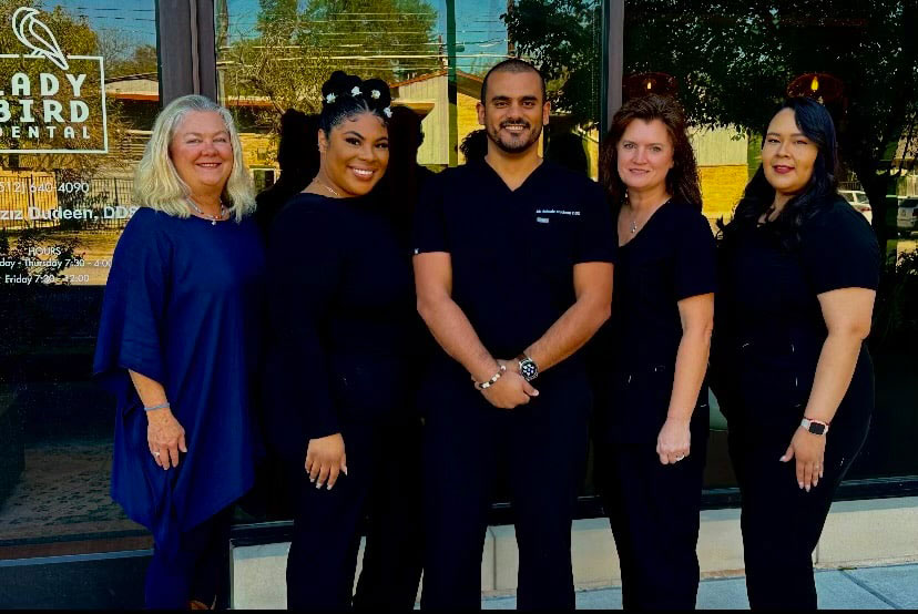 A group of five individuals posing together in front of a salon, with one person standing slightly apart from the rest, all smiling and dressed in professional attire.