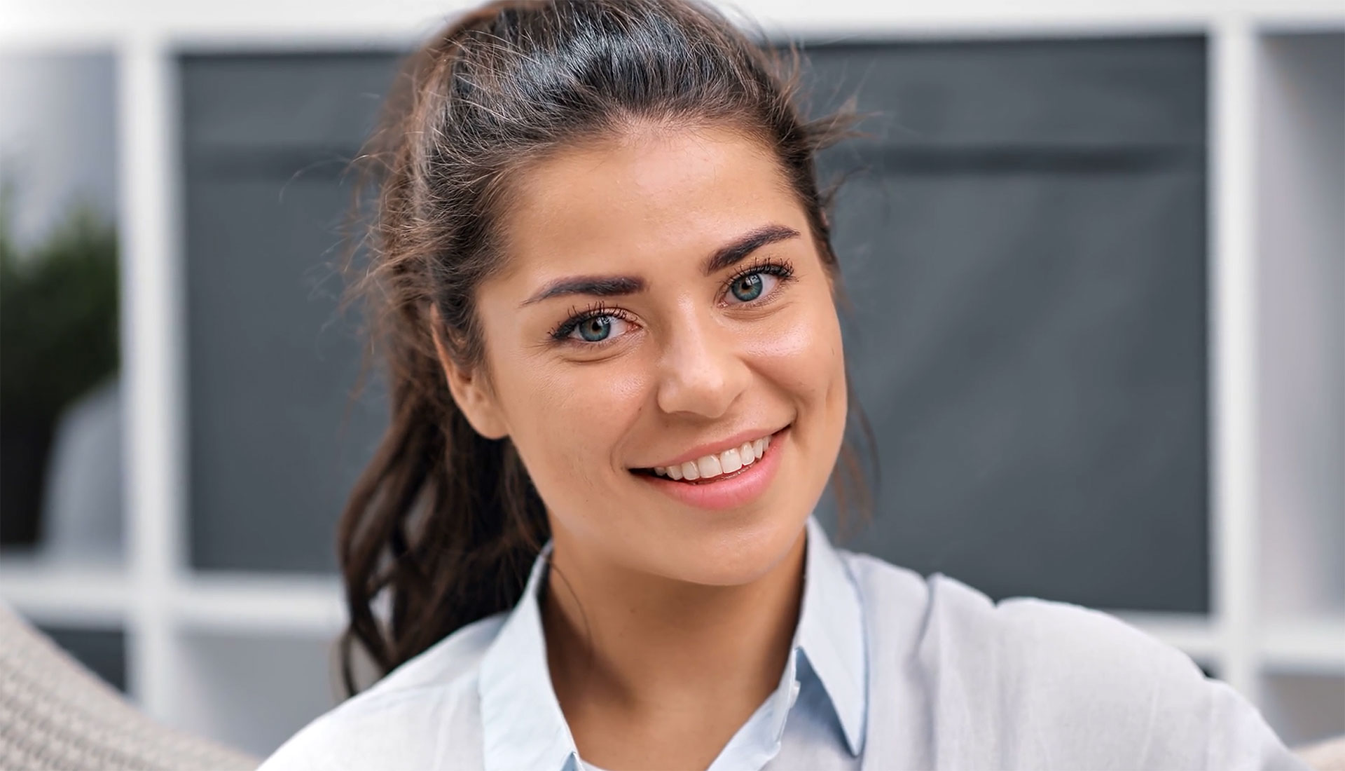 The image shows a woman with a bright smile, looking directly at the camera, wearing a white shirt and a dark blazer, set against an indoor background with neutral tones.