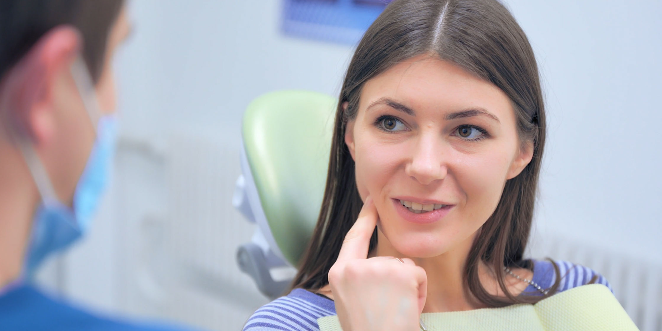 The image shows a woman sitting in a dental chair, receiving dental care from a professional who appears to be conducting an examination or treatment.
