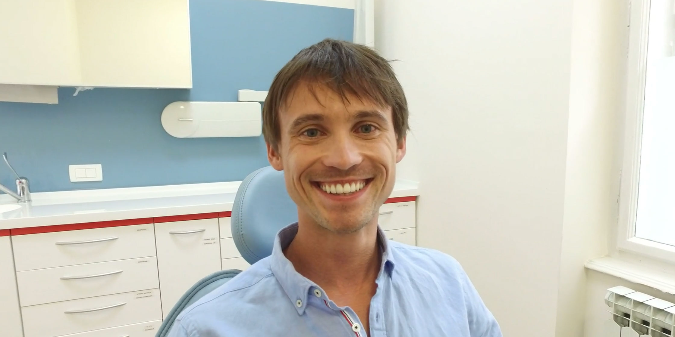 The image shows a smiling man sitting in a dental chair with his mouth open, wearing a blue shirt and glasses, in front of a dental office setting with equipment visible behind him.
