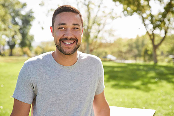 The image shows a man standing outdoors in a park setting, smiling at the camera.