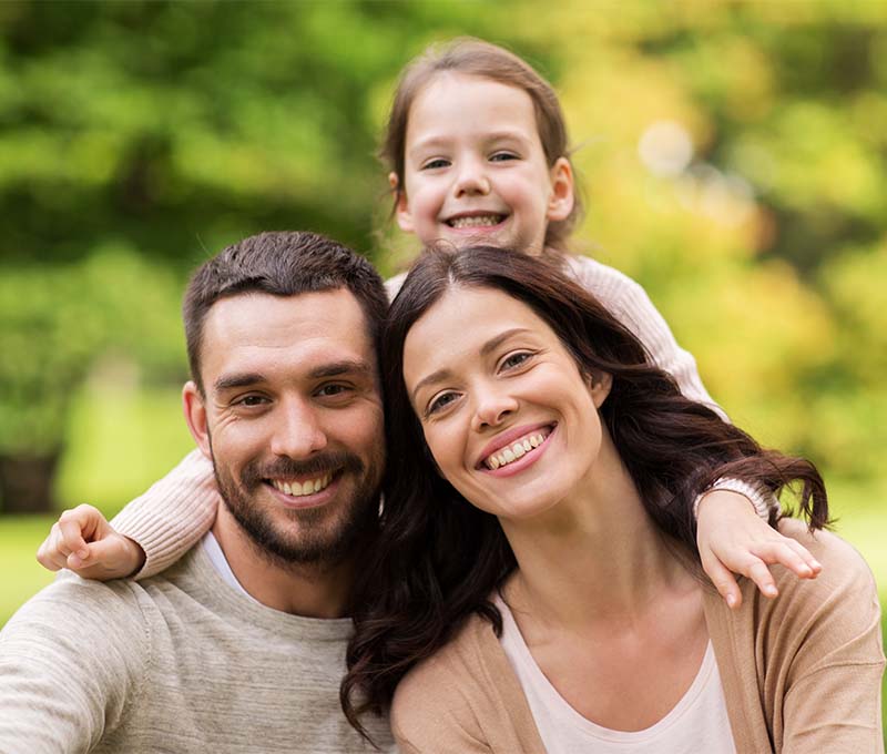 The image shows a family of four - two adults and two children, posing outdoors during daylight, with the adults smiling at the camera and the children looking away.
