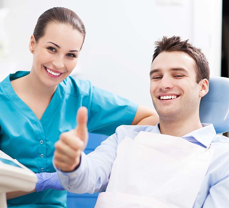 The image shows a dental professional, likely a dentist, giving a thumbs-up sign while standing next to a patient who is seated in a dental chair with a smiling expression.