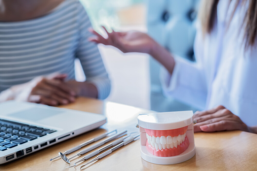 Dentist showing and explaining teeth disease treatment to patient using teeth model denture and explorer mirror tool in dental clinic office. Healthcare concept.