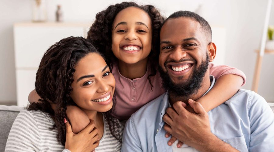 Love And Care. Portrait of cheerful African American family of three people hugging sitting on the sofa at home, posing for photo and looking at camera. Smiling young girl embracing her parents